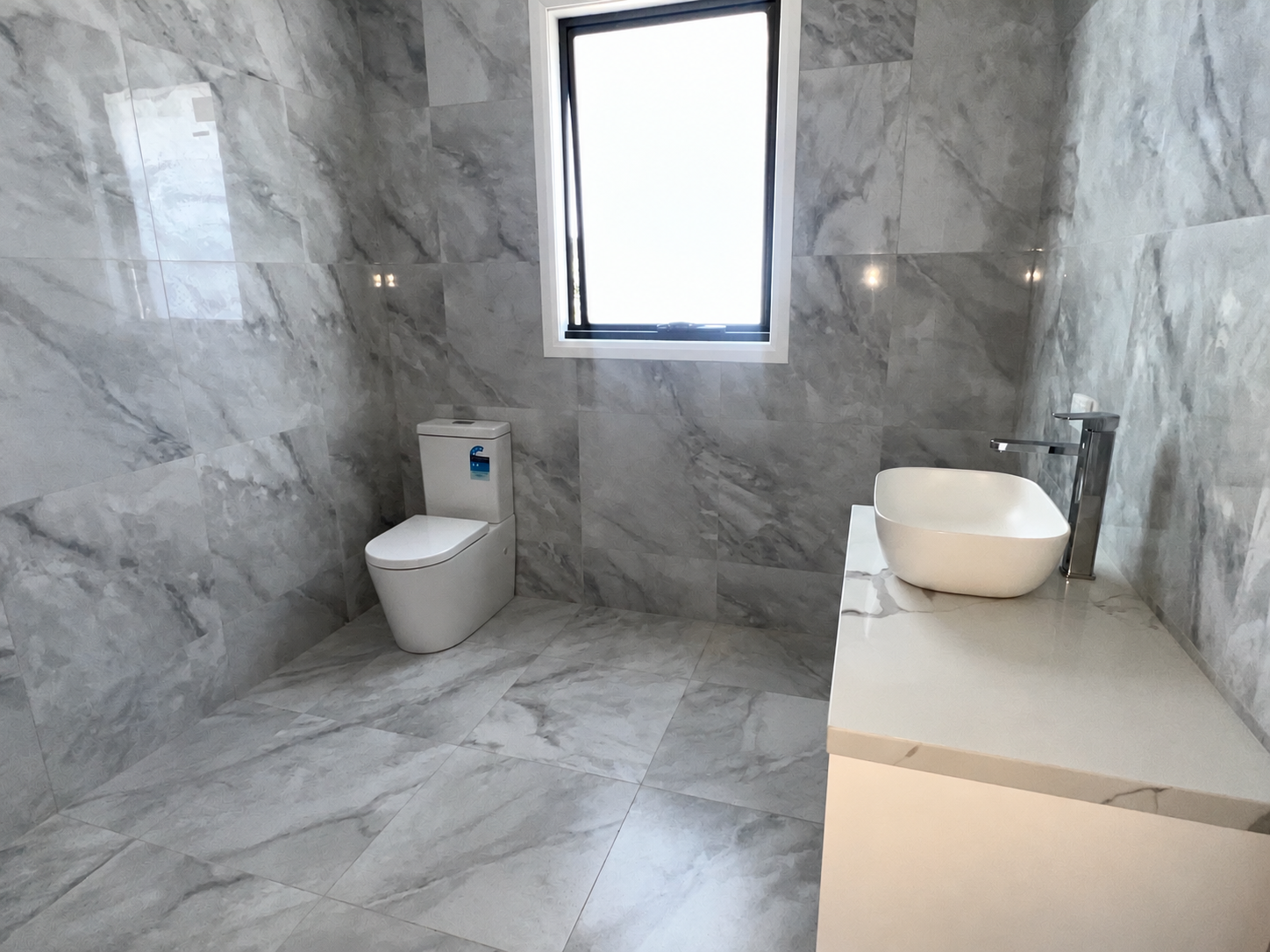 Bathroom fit-off with a white toilet, vessel basin and chrome tap against grey marble-look tiles.