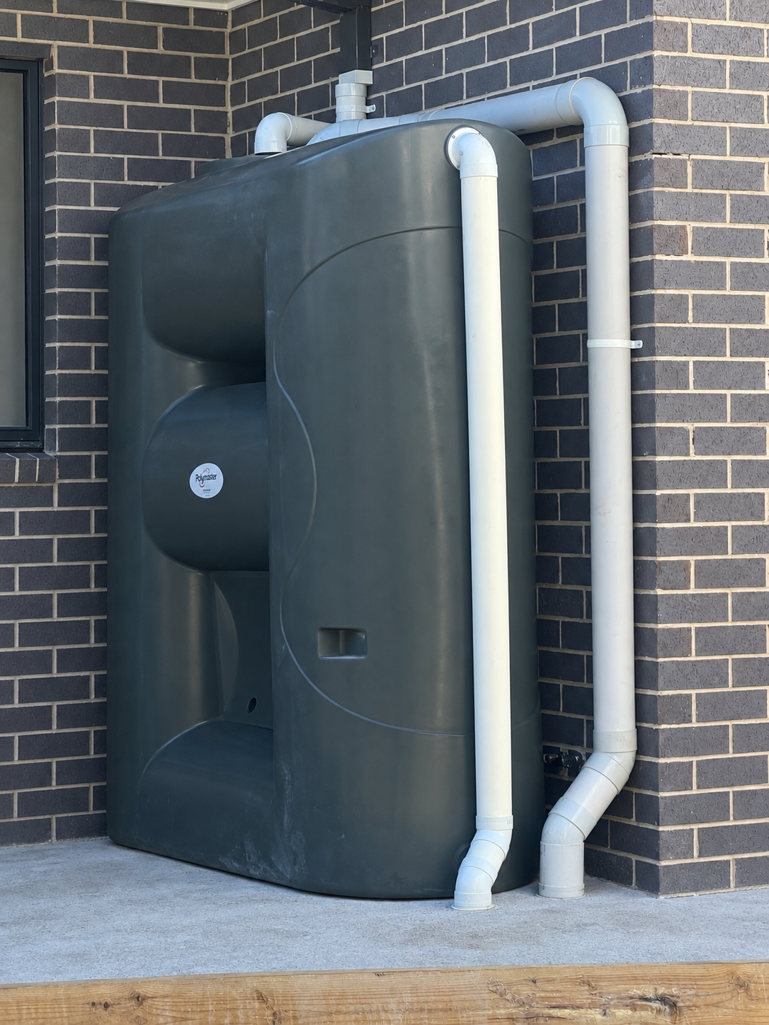Rainwater tank beside a dark brick wall with connected white pipework.